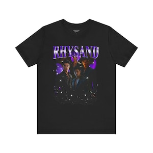 May include: Black t-shirt with a purple graphic that reads "RHYSAND". The design features images of several people and a starry background, giving it a retro aesthetic. The text is in a stylized font.