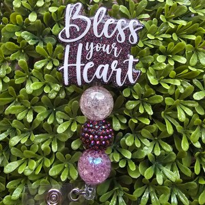May include: A sparkly pink and white glitter badge reel with the words "Bless your Heart" in white lettering. The reel has a clear plastic clip and a silver swivel hook.