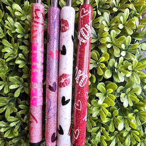 May include: Four glitter pens with silver tips and black tips. The pens are decorated with hearts, kisses, and the word "BE MINE" in pink. The pens are set against a backdrop of green foliage.