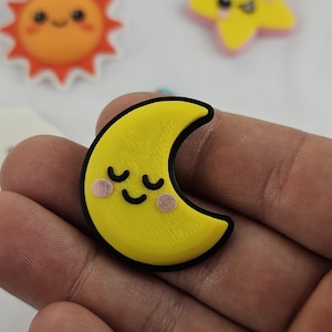 以下が含まれることがあります： 黒い縁取りと閉じられた目を持つ、黄色い三日月型のチャームが手に持たれています。背景には、笑顔の太陽と星など、すべて漫画のような特徴を持つ他のチャームがあります。チャームはおそらく、工芸品やジュエリー用です。
