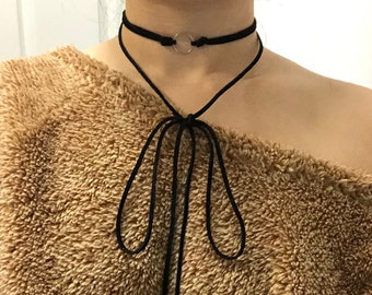 May include: A black choker necklace with a silver circle pendant and a long, thin black cord that ties at the front. The necklace is worn over a brown fuzzy sweater.
