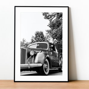May include: Black and white photograph of a vintage car parked in front of a tree. The car is a classic sedan with a chrome bumper and large headlights. The car is in focus, while the background is blurred.