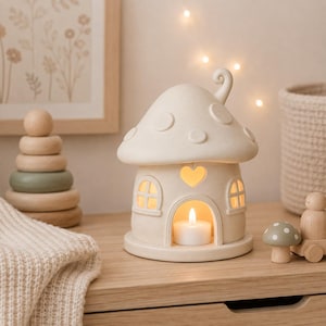 May include: A cream-coloured ceramic mushroom-shaped house candle holder with a rounded roof, a heart-shaped cutout above the arched doorway, and two small arched windows. A white tea light candle glows inside the structure, illuminating the interior.