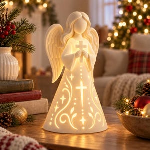 May include: A cream-coloured ceramic angel figurine with an illuminated interior. The angel holds a cross in its hands, and its gown features cut-out designs of a central cross, swirling vine patterns, and small stars that glow from within.