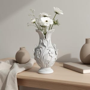 Può includere: Un vaso in ceramica bianca con un motivo floreale e fogliare in rilievo, caratterizzato da un bordo svasato e una base a piedistallo. Il vaso è riempito con fiori bianchi e delicata nebbiolina, con una superficie scolpita e strutturata.