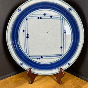 May include: A white ceramic plate with a blue and green geometric design. The plate has a raised rim and a slightly textured surface.