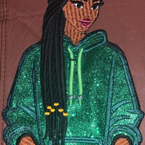 Peut inclure: Patch brodé représentant une femme avec de longues tresses noires portant un sweat à capuche vert pailleté. Le sweat à capuche a un cordon vert et la femme a des perles jaunes dans ses tresses.