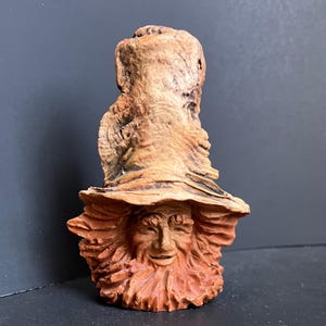 May include: A ceramic sculpture of a whimsical face with a large, textured hat. The face has a long, flowing beard and a friendly expression. The sculpture is painted in shades of brown and orange.
