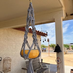 Puede incluir: Un macetero de macramé gris con borla cuelga de un gancho en el techo de un porche. Una maceta de cerámica amarilla está suspendida en el macetero.