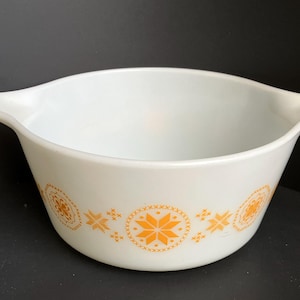 May include: White Pyrex mixing bowl with orange floral and starburst designs. The bowl has two handles and a wide rim. The orange designs are evenly spaced around the bowl's exterior. A vintage kitchenware item.