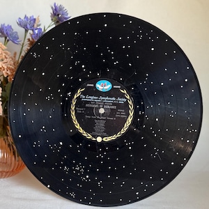 Puede incluir: Un disco de vinilo negro con puntos blancos pintados en él, que se asemeja a un cielo estrellado. La etiqueta del disco presenta el texto "The Longines Symphonette Society" y "Memories of Romance".