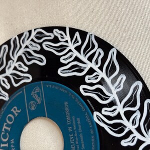 May include: A black vinyl record with a white painted design of a leafy branch. The record label is blue and says "VICTOR" and "BELIEVE IN TOMORROW".