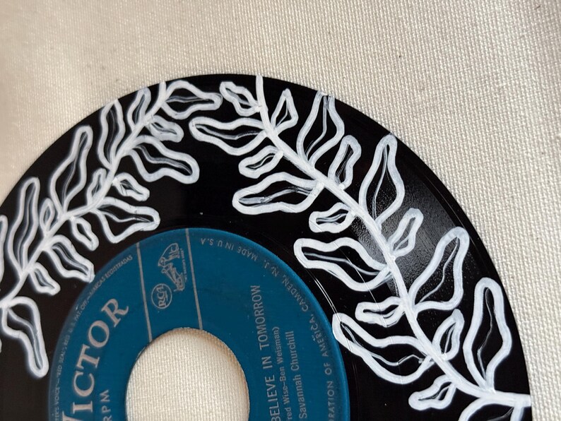 May include: A black vinyl record with a white painted design of a leafy branch. The record label is blue and says "VICTOR" and "BELIEVE IN TOMORROW".