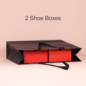 May include: A black paper shopping bag with two red shoe boxes inside. The bag has black handles and the text "2 Shoe Boxes" is above the bag.