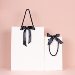 May include: Two white gift bags with black satin ribbon bows and handles. The larger bag has a longer handle and bow. The bags are set against a soft pink background, creating a clean and elegant aesthetic.