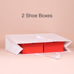 May include: A white paper shopping bag with two red shoe boxes inside. The bag has white handles and the text "2 Shoe Boxes" is printed on the top. The background is a solid light pink.
