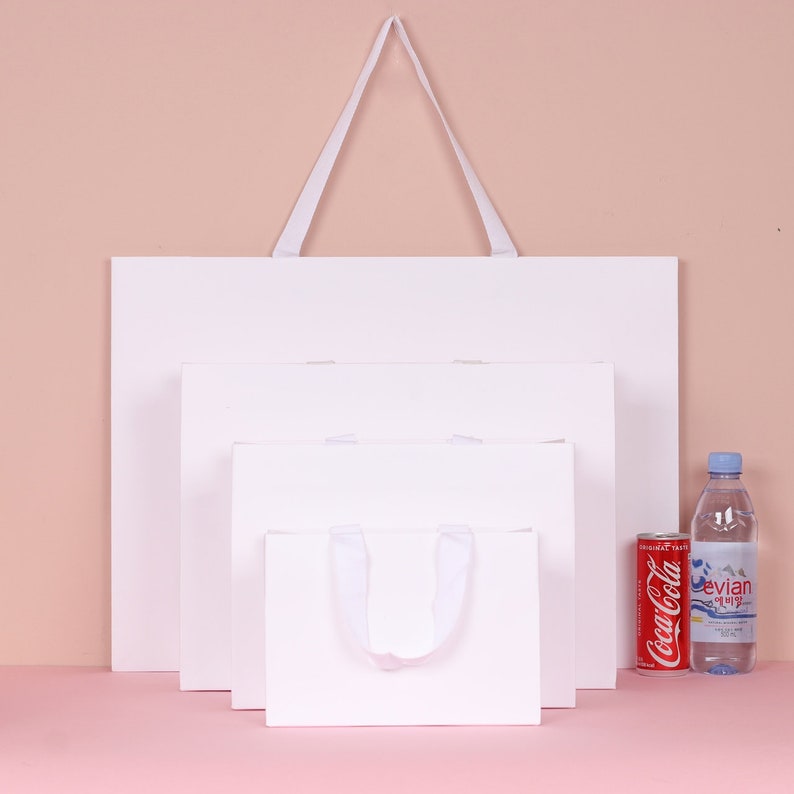 May include: Four white paper shopping bags of varying sizes, with white ribbon handles. A red Coca-Cola can and a bottle of Evian water are next to the bags. The bags are arranged against a light pink background.