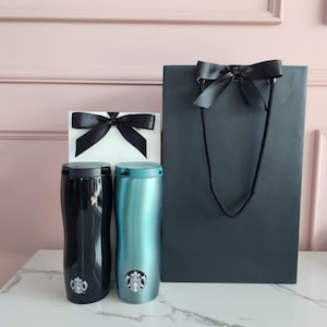 May include: Two insulated travel mugs, one black and one teal, with the Starbucks logo, sit next to a white gift box and a dark gray shopping bag, all adorned with black satin bows. The items are arranged on a white marble surface.