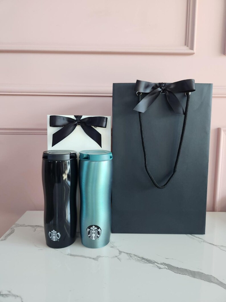 May include: Two insulated travel mugs, one black and one teal, with the Starbucks logo, sit next to a white gift box and a dark gray shopping bag, all adorned with black satin bows. The items are arranged on a white marble surface.