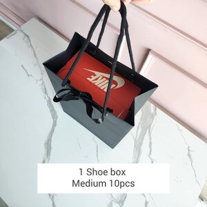 May include: A black paper shopping bag with black rope handles holds a red shoe box with a white Nike logo. The bag is tied with a black ribbon. The text on the bottom reads: "1 Shoe box Medium 10pcs."