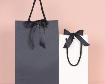 May include: Two gift bags, one charcoal gray and one white, are displayed against a pink background. Both bags have black ribbon bows and matching handles. The larger bag is rectangular, while the smaller one is more square.