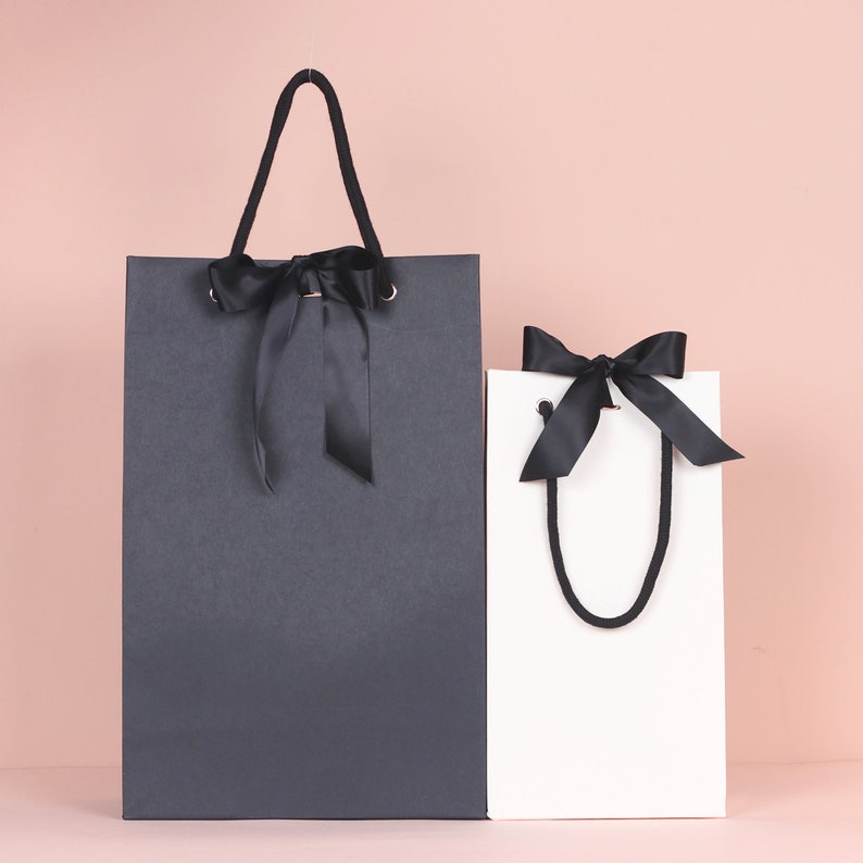 May include: Two gift bags, one charcoal gray and one white, are displayed against a pink background. Both bags have black ribbon bows and matching handles. The larger bag is rectangular, while the smaller one is more square.