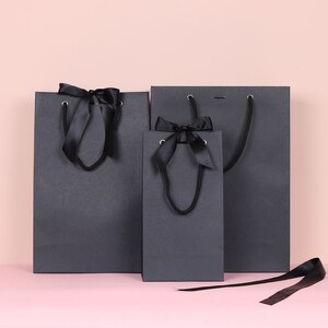 May include: Three dark gray gift bags of varying sizes, each with black satin ribbon handles and bows. The bags are arranged against a pink background, with one ribbon lying loose.