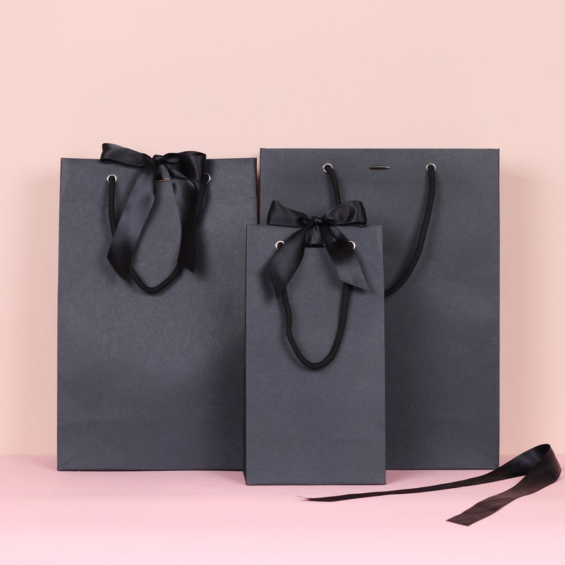 May include: Three dark gray gift bags of varying sizes, each with black satin ribbon handles and bows. The bags are arranged against a pink background, with one ribbon lying loose.
