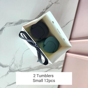 May include: A white gift bag tied with a black ribbon, containing two tumblers. One tumbler is black, and the other is teal. The text on the bag reads "2 Tumblers Small 12pcs."