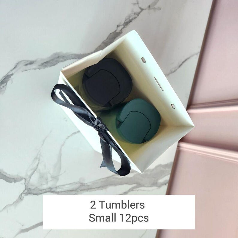 May include: A white gift bag tied with a black ribbon, containing two tumblers. One tumbler is black, and the other is teal. The text on the bag reads "2 Tumblers Small 12pcs."