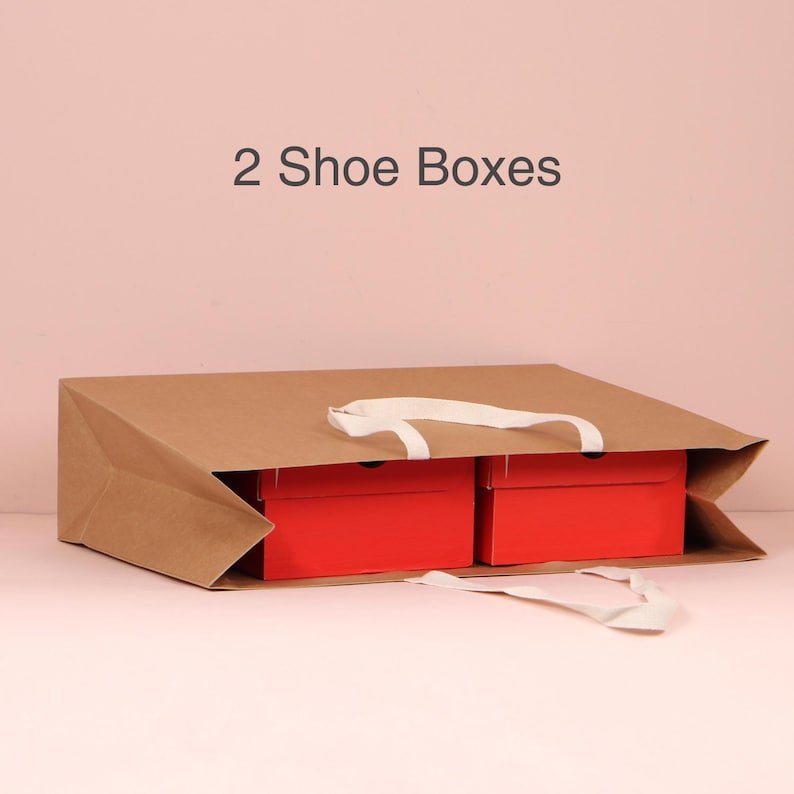 May include: A brown paper shopping bag with white handles, containing two red shoe boxes. The text "2 Shoe Boxes" is displayed above the bag against a pink background.