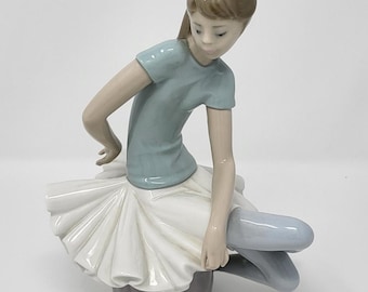 May include: A porcelain figurine of a young girl dressed in a blue shirt and white tutu, sitting on a gray base. She is holding her leg with one hand and her other hand is resting on her knee.