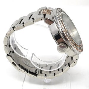 May include: A silver-toned wristwatch with a stainless steel band. The watch face is surrounded by a ring of small, sparkling stones. The band is made of interlocking links. The watch is displayed on a clear stand.