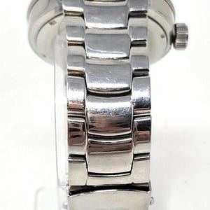 May include: A silver-toned wristwatch with a stainless steel band. The watch has a round face and a textured bezel. The band is composed of rectangular links. The watch is displayed on a clear stand.