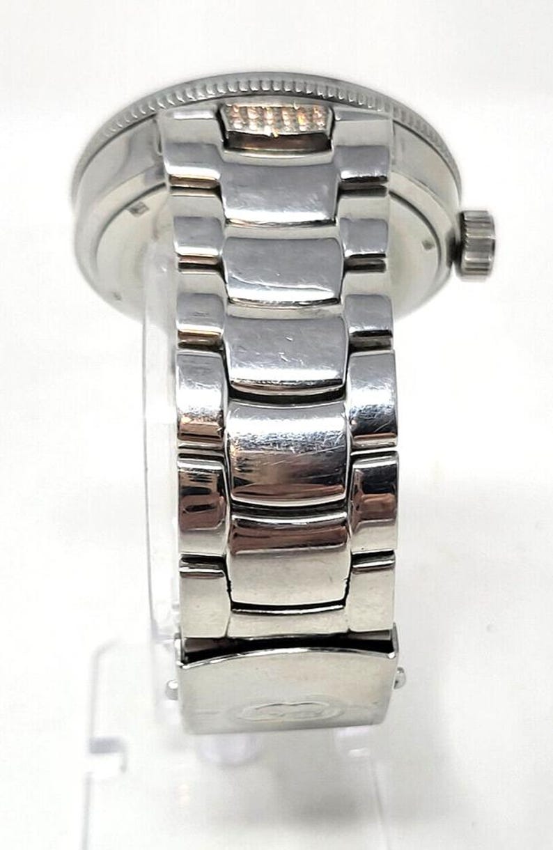 May include: A silver-toned wristwatch with a stainless steel band. The watch has a round face and a textured bezel. The band is composed of rectangular links. The watch is displayed on a clear stand.