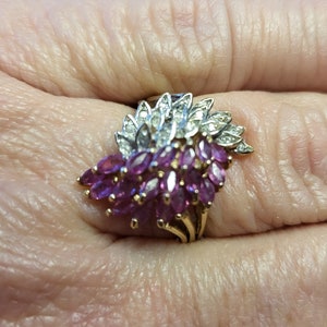 May include: A gold ring featuring a cluster of marquise-cut, deep pink gemstones, accented with smaller clear stones. The ring has a floral design, with the gemstones arranged to resemble petals. The ring is set against a skin-toned background.