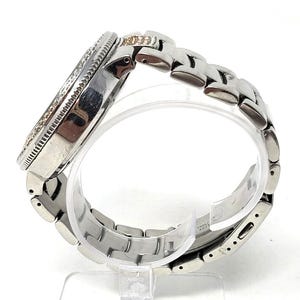 May include: A silver-toned wristwatch with a round face and a linked band. The watch face is encircled with small, clear stones. The band is made of interlocking metal links. The watch is displayed on a clear stand.