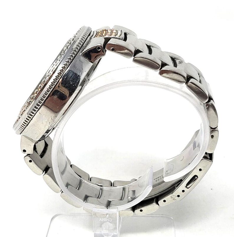 May include: A silver-toned wristwatch with a round face and a linked band. The watch face is encircled with small, clear stones. The band is made of interlocking metal links. The watch is displayed on a clear stand.