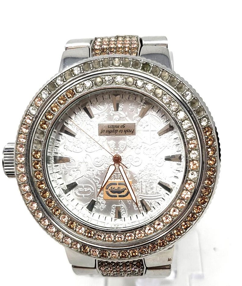 May include: A silver-toned wristwatch with a round face, adorned with clear and brown crystals around the bezel and on the band. The watch face has a patterned design and the text "Fresh to depths of 50 meters".