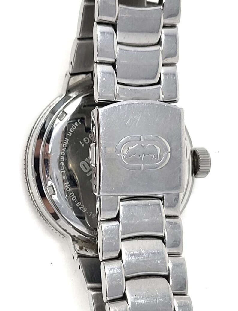May include: Close-up of a silver-toned wristwatch with a metal band. The watch face is visible, with the words "Japan movement" and "G1" printed on it. The clasp features a logo.