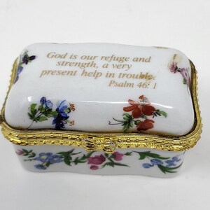 May include: A white porcelain trinket box with a gold trim and floral design. The lid features the text "God is our refuge and strength, a very present help in trouble. Psalm 46:1".