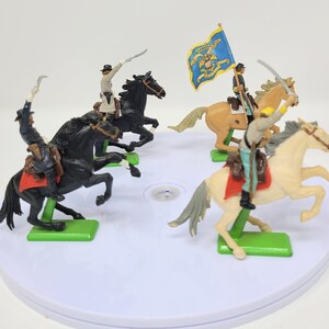 May include: Four toy cavalry soldiers on horseback, each with a different colored horse and uniform. The soldiers are holding swords and are posed in a battle formation. The horses are all standing on a white circular base.