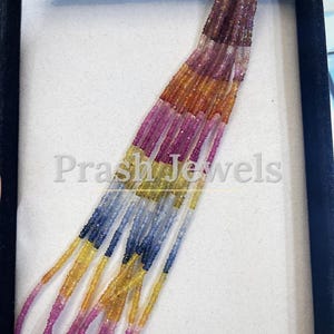 May include: A strand of faceted, multi-colored gemstone beads arranged in a gradient pattern, transitioning from deep pink and orange to yellow, green, and blue. The beads are strung on thin wire, and the text "Prash Jewels" is visible in the center.