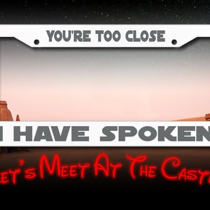May include: White metal license plate frame with the text "You're Too Close. I Have Spoken. Let's Meet At The Castle" in black and red lettering. The frame is set against a desert landscape.