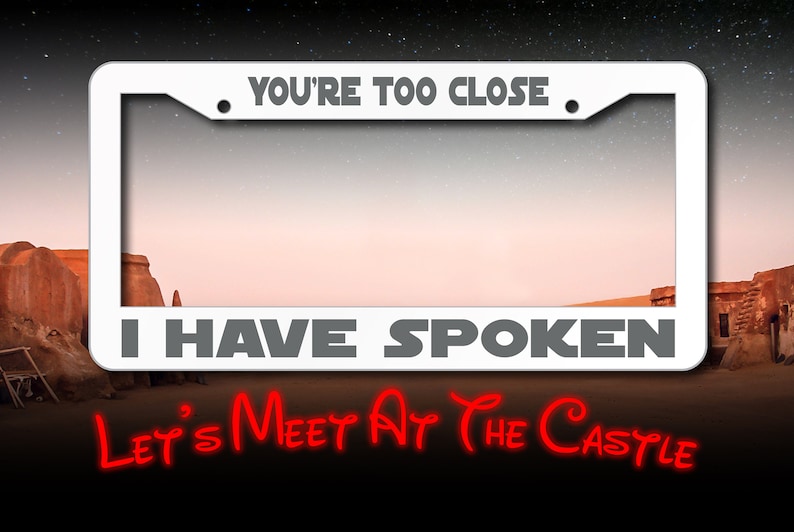 May include: White metal license plate frame with the text "You're Too Close. I Have Spoken. Let's Meet At The Castle" in black and red lettering. The frame is set against a desert landscape.