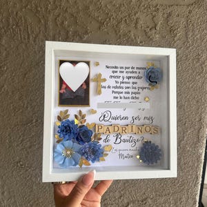 Puede incluir: Una caja de sombra con marco blanco con flores de papel azul, detalles dorados y texto en español. La caja contiene una foto con un corazón encima, una cruz de madera y la pregunta "¿Quieren ser mis Padrinos de Bautizo?"