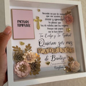 May include: A white shadow box frame containing a pink picture template, a gold cross, and paper flowers in shades of pink, beige, and gold. The text reads: Necesito un par de manos que me ayuden a crecer y aprender. Tia Evelyn y Tio Gabriel Quieren ser mis PADRINOS de Bautizo?