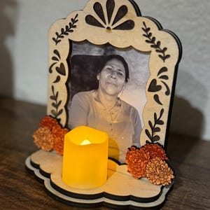 May include: A wooden Day of the Dead memorial with a black and white photo of a woman. The frame is decorated with laser-cut floral designs and orange and yellow flowers. A battery-operated candle sits in front of the photo.