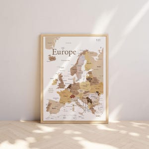 May include: A framed map of Europe with a light wood frame. The map is in shades of brown and beige, with country names labeled. The word "Europe" is at the top. The map is leaning against a white wall.