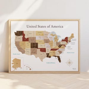 以下が含まれることがあります： 額縁に入ったアメリカ合衆国の木製地図。地図には、茶色とベージュの色合いの個々の州が表示され、州名はセリフ体で印刷されています。地図は白い背景にあり、太平洋と大西洋が含まれています。